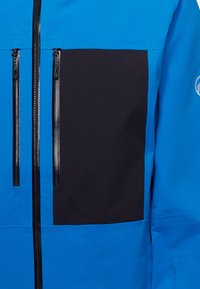 Blue waterproof jacket featuring a large black patch, two black zippered pockets, and contrasting texture on the chest area.