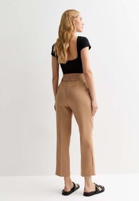 Top noir à manches courtes avec découpe dans le dos, associé à un pantalon taillé marron clair avec passants de ceinture et poches structurées. Porté avec des sandales noires.