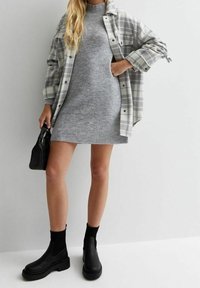 Femme portant une robe en maille côtel ée grise, une veste-chemise à manches longues à carreaux, des bottines noires et tenant un petit sac à main noir.