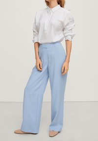 Pantalons larges bleu clair en tissu doux, associés à une chemise blanche à boutons avec un col rabattu et des manches retroussées.