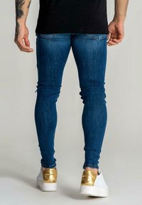 Těsné džíny ve tmavě modrém denimu s přiléhavým střihem a mírným vyblednutím. Párek bílých bot s kovovými zlatými akcenty na patě.