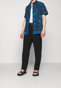 Blauwe shirt met korte mouwen en zwart bladpatroon, zwarte broek en zwarte sandalen. Heeft een gebrandmerkte taille en een ontspannen pasvorm.