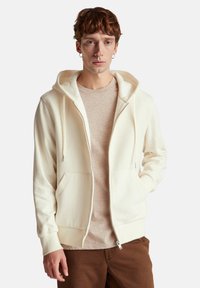 Sudadera blanca con cremallera y textura acanalada en el frente, con cordones y bolsillos delanteros, usada sobre una camiseta beige claro y pantalones marrones.