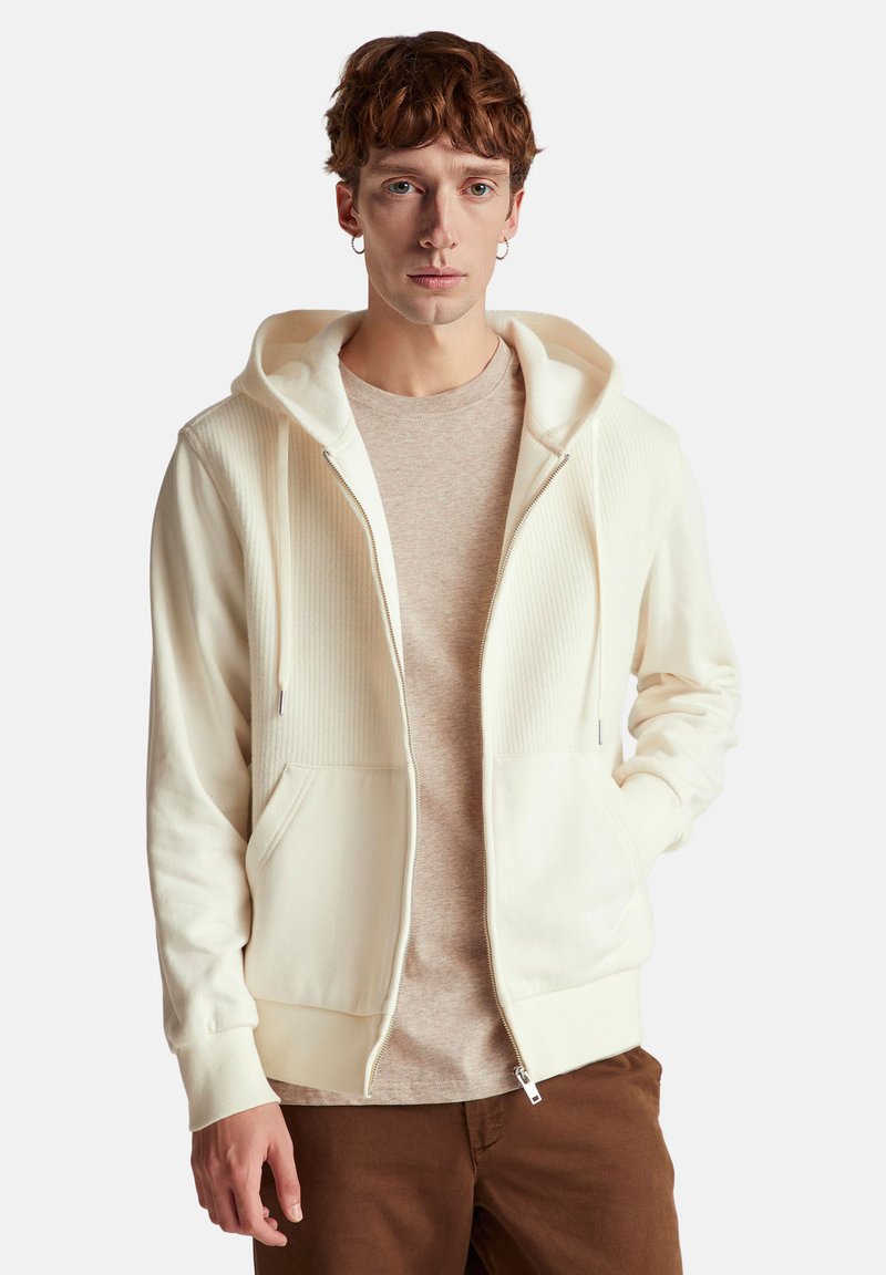 Bílá mikina na zip s řasenou strukturou na přední straně, se šňůrkami a předními kapsami, nošená přes světle béžovou košili a hnědé kalhoty.