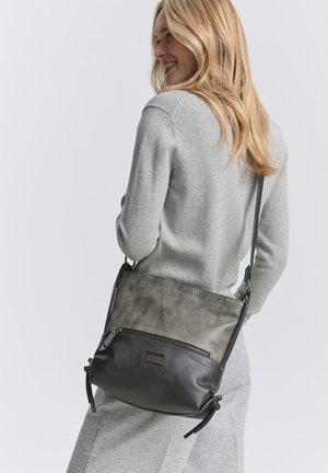 Kvinde med langt blondt hår iført en lysegrå sweater og nederdel, bærende en tofarvet skuldertaske med sorte stropper og lynlåslomme.