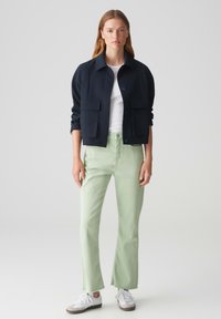 Giacca cropped blu navy con grandi tasche frontali, abbinata a pantaloni a zampa di elefante verde menta e sneaker bianche, su uno sfondo neutro.
