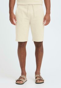 Shorts en maille beige avec taille élastique et cordon de serrage. Tissu texturé et poches latérales. Portés avec des sandales marron.