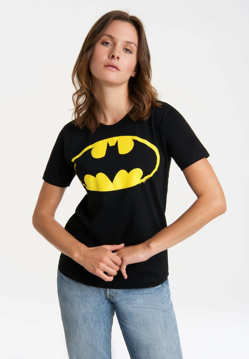 Young woman with wavy brown hair wearing a black Batman logo t-shirt and light blue jeans, standing against a plain white background.