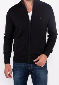 Černý svetr na zip vyrobený z měkké látky, s vysokým límcem a malým logem na levé hrudi, v kombinaci s modrými džínami.