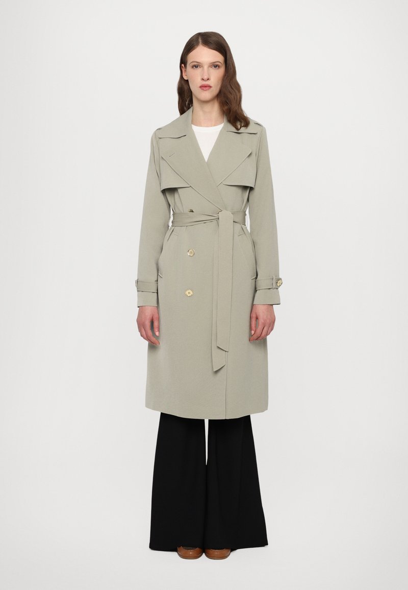 Manteau trench gris clair avec des boutons dorés à double boutonnage, taille ceinturée et larges revers, associé à un pantalon noir à jambes larges.