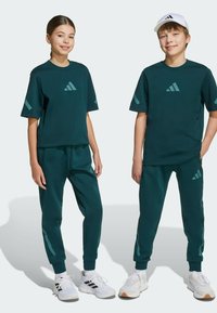Conjunto deportivo verde oscuro con camiseta de manga corta y pantalones ajustados. Presenta el logo de Adidas y acentos de contraste. Zapatillas blancas complementan el look.