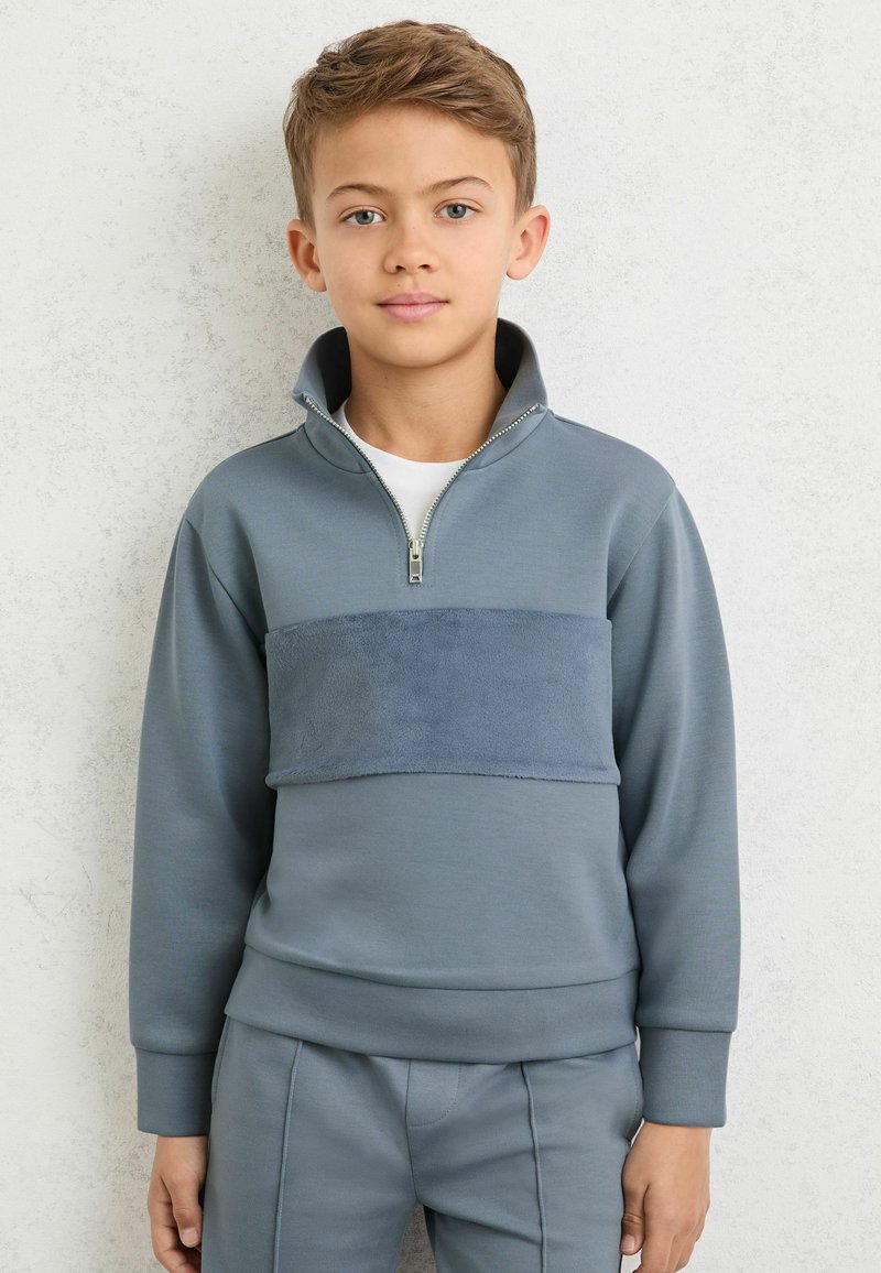 Sweatshirt gris avec un col à zip et une bande en tissu pelucheux traversant la poitrine, associé à un pantalon gris assorti. Texture lisse, design décontracté.