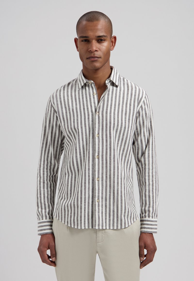 Homme portant une chemise à rayures verticales grises et blanches boutonnée, avec un pantalon beige, sur un fond clair uni.