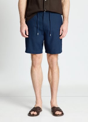 Man wearing navy blue drawstring shorts, brown woven sandals, and a dark brown buttoned shirt standing against a white background.