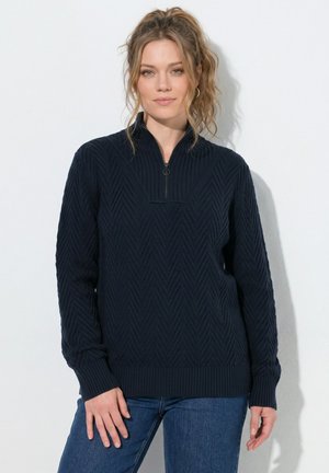 Kvinde med lyst brunt hår i en høj hestehale iført en mørk marineblå tekstureret sweater med lynlås ved halsen og blå jeans, stående mod en hvid baggrund.