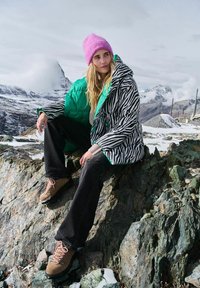 Žena v ružovej čiapke a kabáte s pruhovaným zebrovaním sedí na skalnatom teréne s zasneženými horami a zamračenou oblohou v pozadí.