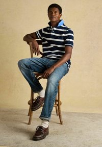 Homme assis sur une chaise en bois portant un polo rayé bleu marine et blanc, un jean bleu, des chaussettes blanches et des chaussures bateau en cuir marron, contre un mur beige.