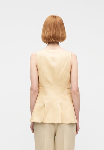 Γυναίκα με κοντά κόκκινα μαλλιά που φοράει μπεζ αμάνικο ραμμένο τοπ και ταιριαστό παντελόνι, στραμμένη προς τα πίσω μπροστά σε απλό ανοιχτό φόντο.