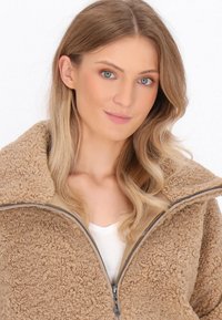 Giacca in pile morbido e fluffy color tan con colletto alto, chiusura a zip e fodera liscia. Presenta una superficie testurizzata e accenti beige a contrasto.