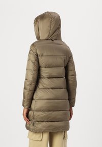Persoon in een lange, olijfgroene gewatteerde jas met een capuchon, gezien vanaf de achterkant tegen een eenvoudige achtergrond.