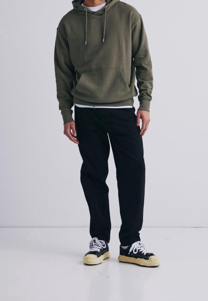 Homme portant un sweat à capuche vert olive, un pantalon noir et des baskets noires avec des lacets blancs, debout sur un fond clair uni.