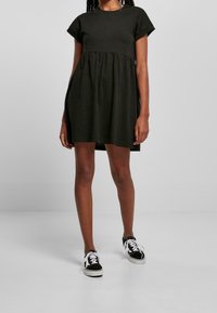 Černé šaty s krátkým rukávem a rozšířenou sukni, se stahovaným pasem. Kombinovány s černobílými teniskami, stojící na světlém pozadí.