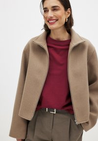 Beige wool-blend jacket with a zip front, high collar, and relaxed fit, worn over a maroon shirt, paired with gray trousers and a black belt.