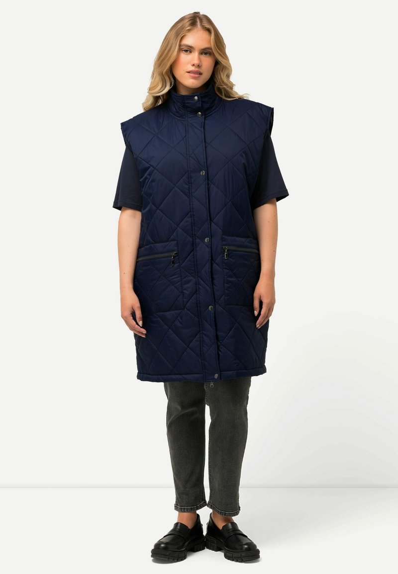 Gilet trapuntato blu navy con colletto alto, dotato di tasche laterali con zip, bottoni a scatto e una lunghezza fino al ginocchio. Indossato sopra una camicia scura e jeans.