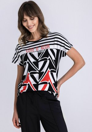 Vrouw draagt een zwart-wit-rood gestreept T-shirt met de tekst "CÔTE D'AZUR SAINT TROPEZ", gecombineerd met een zwarte broek, tegen een effen achtergrond.