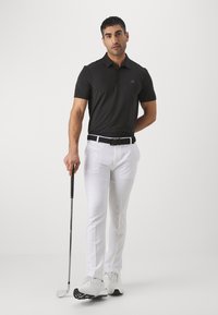 Polo preta com gola e mangas curtas, combinada com calças brancas e um cinto preto. O indivíduo segura um taco de golfe.