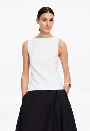 Frau mit zurückgekämmtem Haar, die ein ärmelloses weißes Oberteil und einen schwarzen Rock trägt, Hände in den Taschen, silbernes Armband am linken Handgelenk.