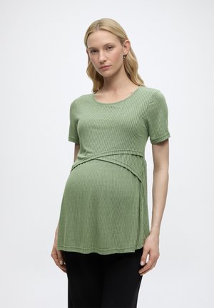 Femme enceinte aux cheveux blonds portant un haut de maternité côtelé vert clair à manches courtes et un pantalon noir sur fond uni.