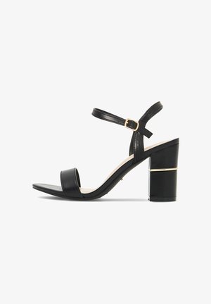 Sandalia negra de punta abierta con tacón cuadrado, correa en el tobillo y hebilla dorada con detalle de adorno dorado en el tacón.