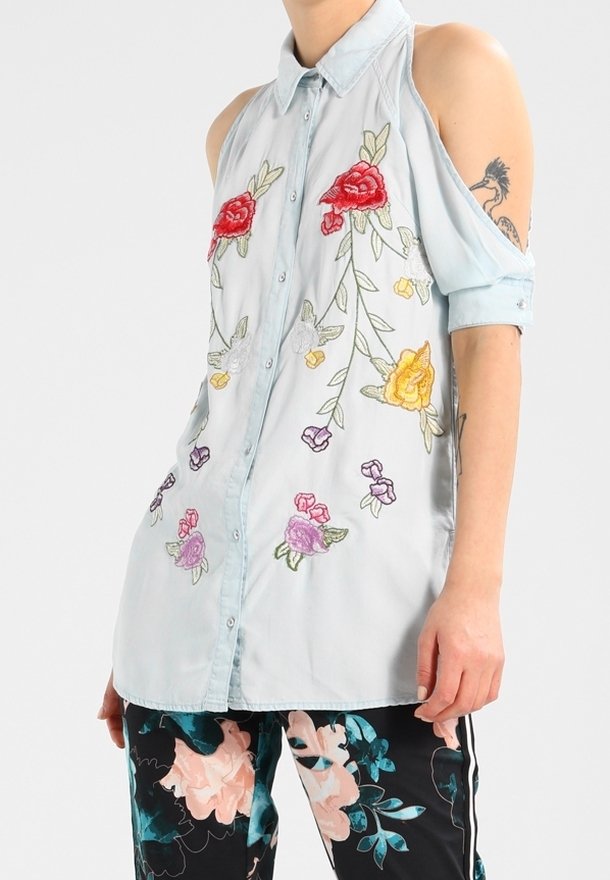 Camicia button-up azzurro chiaro con ricami floreali in rosso, rosa, giallo e viola. Design con spalle scoperte e maniche arrotolate. Tessuto morbido.