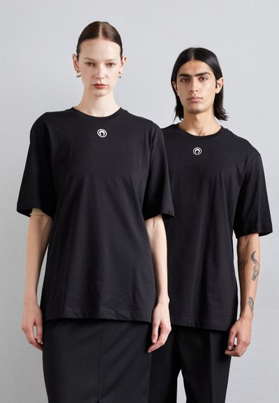 Schwarze, übergroße T-Shirts aus Baumwolle mit rundem Halsausschnitt und kurzen Ärmeln. Ein kleines, rundes Logo ist zentral auf der Brust platziert.