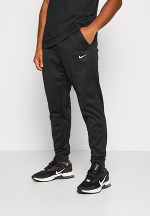Man draagt zwarte Nike joggers en zwarte Nike sneakers, staat met één hand in de zak van de joggers tegen een effen achtergrond.
