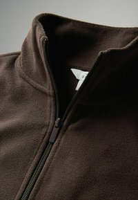 Hnedá fleecová bunda s vysokým golierom a zipsom, ktorá má jemnú textúru a viditeľné detaile šitia. Vo vnútri je pripevnený štítok s pokynmi na údržbu.