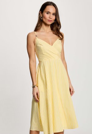 Donna che indossa un vestito midi senza maniche a righe gialle e bianche con scollo a V, accessoriato con braccialetti e orecchini dorati.