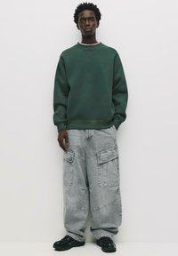 Zelená mikina, šedé cargo kalhoty s velkými kapsami a černé tenisky. Tkanina vypadá měkce a design je oversize a uvolněný.
