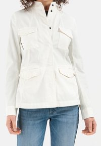 Biała kurtka użytkowa z zamkiem błyskawicznym z przodu, wysokim kołnierzem i wieloma kieszeniami. Materiał jest gładki z delikatnym połyskiem.