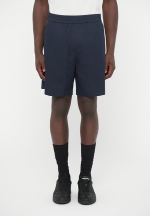 Σκούρα μπλε σορτς από ελαφρύ ύφασμα με ελαστική μέση, σετ με μαύρες κάλτσες έως τον αστράγαλο και μαύρα αθλητικά παπούτσια.