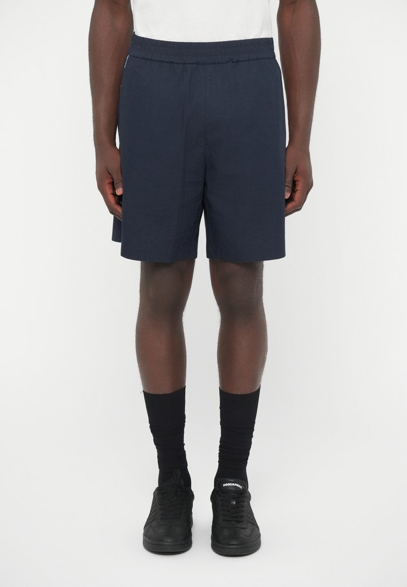 Σκούρα μπλε σορτς από ελαφρύ ύφασμα με ελαστική μέση, σετ με μαύρες κάλτσες έως τον αστράγαλο και μαύρα αθλητικά παπούτσια.