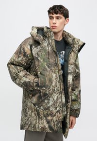 Parka imbottito con motivo mimetico marrone e verde corteccia, caratterizzato da un colletto alto, ampie tasche e chiusura frontale con zip.