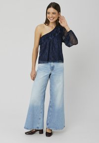 Haut asymétrique bleu marine avec des volants, associé à un jean large bleu clair et des chaussures sombres et brillantes, le tout sur un fond neutre.