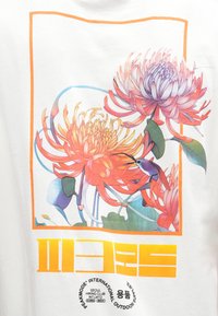 Camiseta blanca con un gráfico floral colorido que presenta flores rojas y moradas dentro de un borde rectangular naranja, y texto en negrita en la parte inferior.