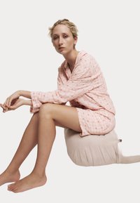 Ljusrosa gingham-pyjamas set med kort ärm och shorts; sitter på en beige, plyschkudde med svansdetalj. Mjuk textur.