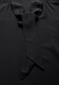 Schwarze Bluse mit einer breiten Schleife am Kragen. Glattes, mattes Material mit einer weichen Textur und ohne sichtbare Muster oder Akzente.
