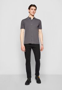 Pilkas trumpomis rankovėmis marškiniai su apykakle, dėvimi su juodomis džinsomis ir juodais batais. Audinys atrodo lygaus, su prigludusia forma.