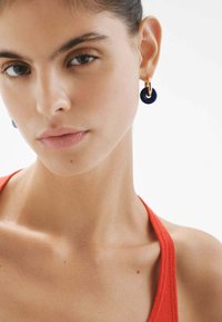 Pendientes de aro dorados con acentos circulares de azul profundo. Textura suave con acabado brillante. Llevados por una modelo con una blusa roja, primer plano de su rostro.