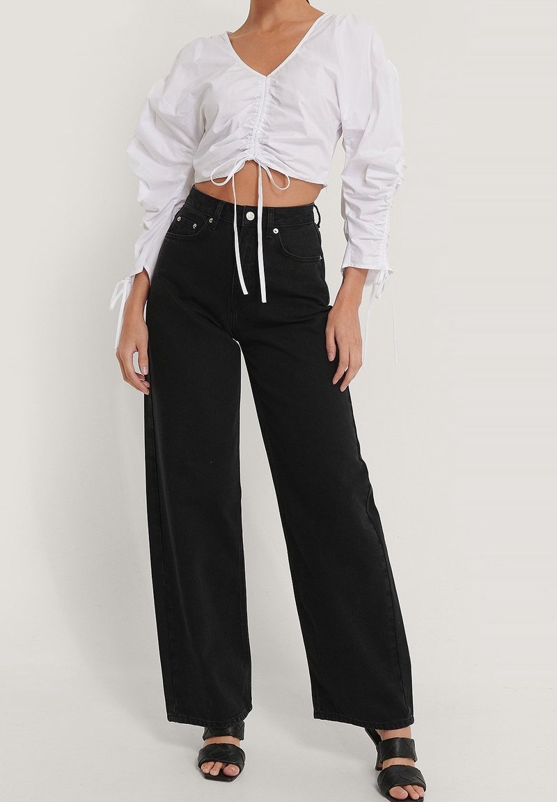 Femme portant un chemisier court blanc froncé à manches longues, un jean noir taille haute à jambes larges, et des sandales noires à bout ouvert.